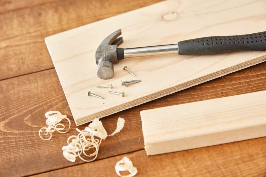 A hammer, nails, and wood shavings resting on a board next to woodworking joints preparation.